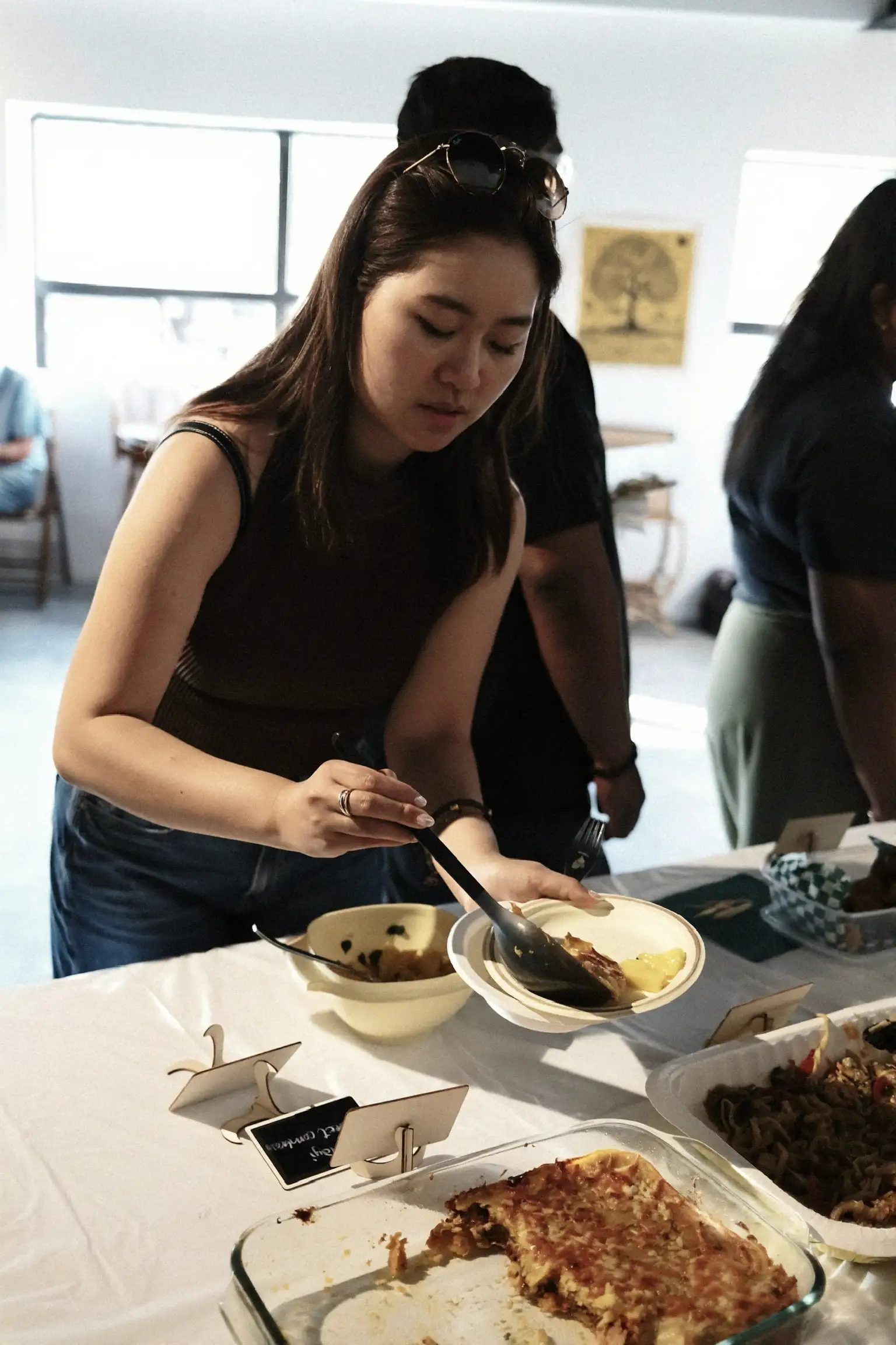 A guest spooning food onto her plate at Sauté Sundays, beside a labelled bake and a tray of noodles