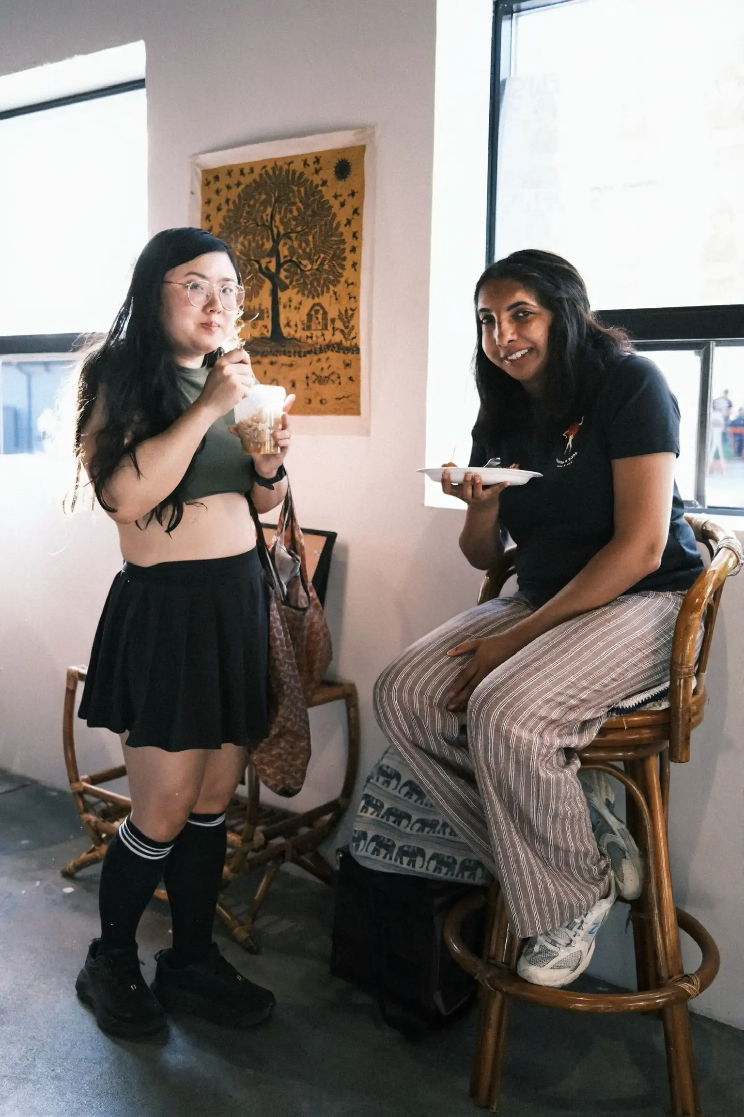 Two guests eating and talking at Sauté Sundays, one perched on a rattan stool with a plate balanced on her hand
