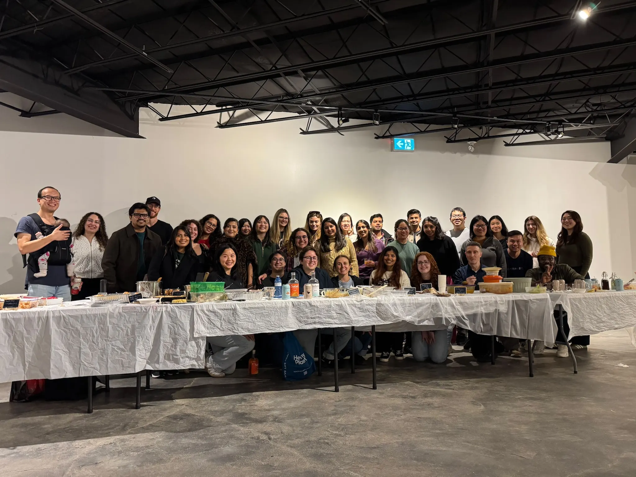 Everyone at Sauté Sundays Friendsgiving 2026 gathered for a group photo behind the spread of dishes at Jrew's in Toronto, one of the night's two babies in a carrier on the left.