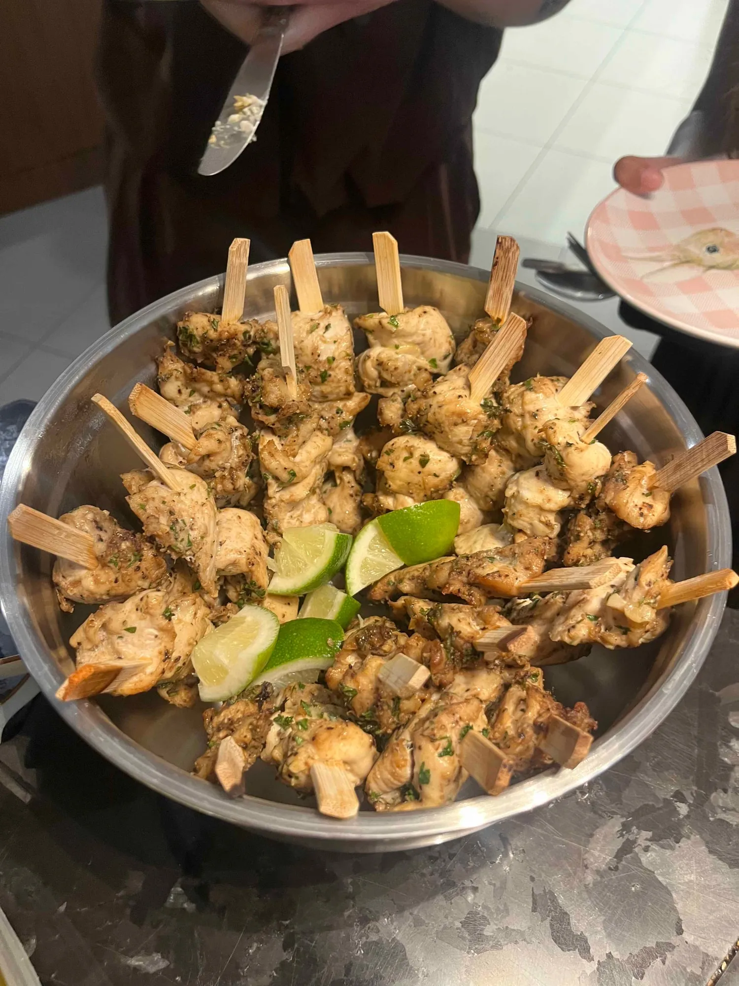 Chicken skewers in a metal bowl with lime wedges, cooked at home and carried in to a Sauté Sundays cookbook club night