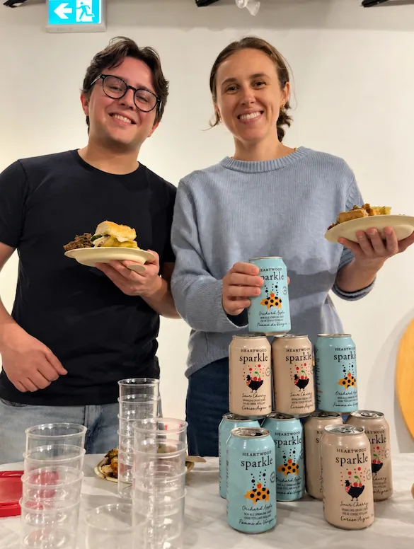 Two guests holding plates of food and cans of Heartwood Sparkle beside a stack of cans and cups at the mocktail bar