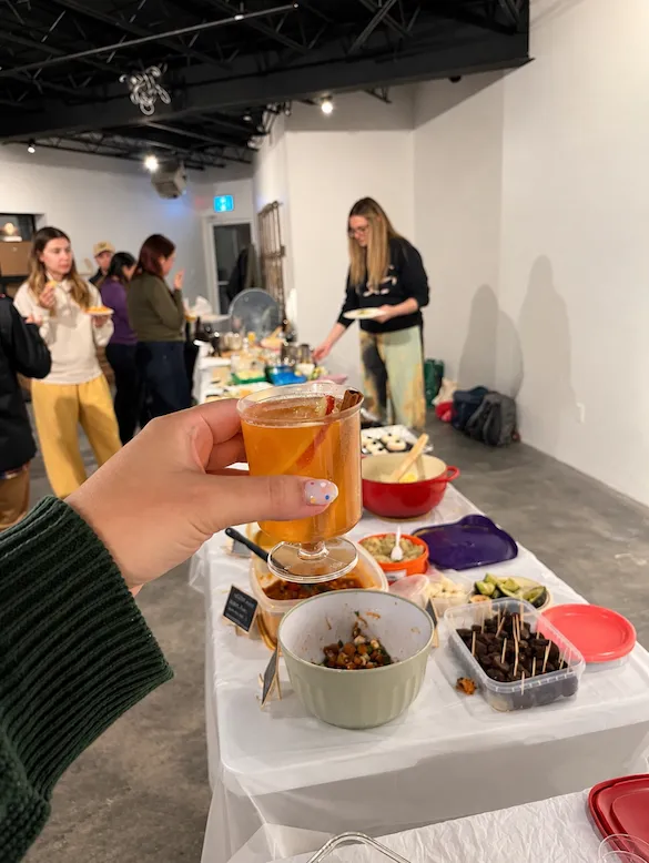 An amber mocktail held up in front of the potluck spread, with guests serving themselves in the background