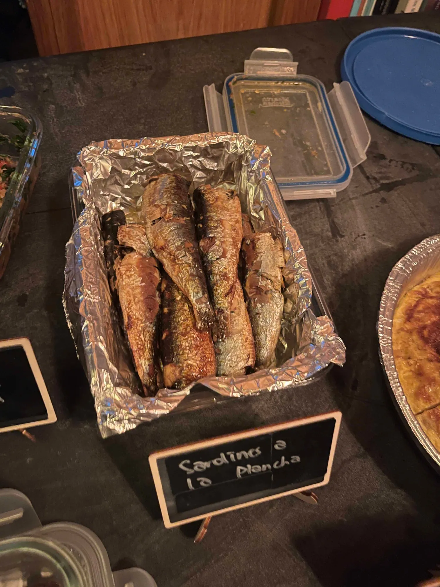Sardines a la plancha in a foil tray with a small chalkboard label naming the dish, among other containers on the table